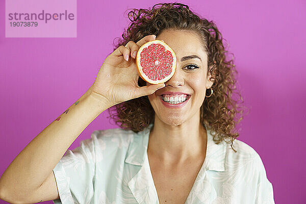 Glückliche Frau hält Grapefruit vor magentafarbenem Hintergrund übers Auge
