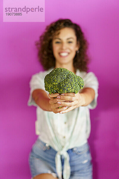 Glückliche Frau hält Brokkoli in der Hand und steht vor magentafarbenem Hintergrund
