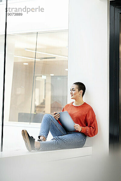 Entspannte Geschäftsfrau sitzt auf der Fensterbank im Bürogebäude und nutzt ein digitales Tablet
