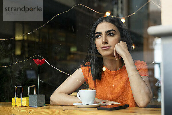 Nachdenkliche junge Frau sitzt mit Kaffee und Smartphone am Tisch im Café