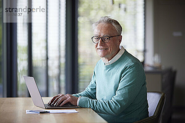 Porträt eines lächelnden älteren Mannes  der zu Hause am Tisch einen Laptop benutzt