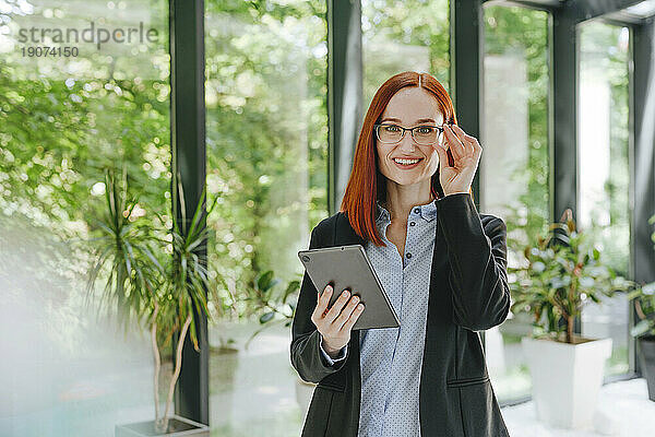 Glückliche rothaarige Geschäftsfrau steht mit Tablet-PC im Büro