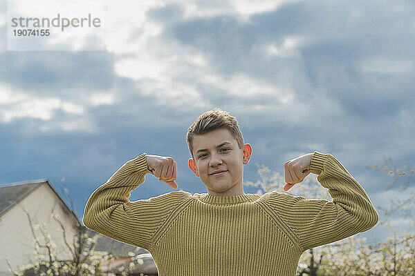 Smiling boy flexing muscles under cloudy sky
