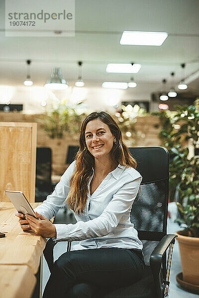 Glückliche junge Geschäftsfrau sitzt mit Tablet-PC auf einem Stuhl am Schreibtisch im Büro