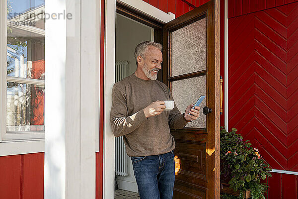 Lächelnder Mann benutzt Smartphone und trinkt Kaffee  der in der Nähe der Tür steht