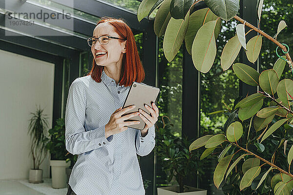 Fröhliche Geschäftsfrau steht mit Tablet-PC neben der Pflanze im Büro