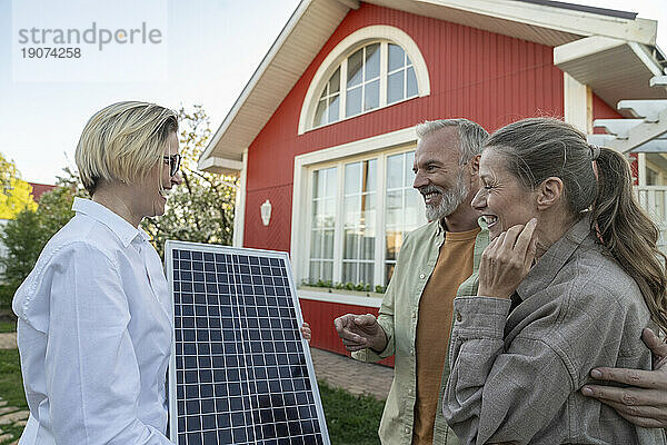 Glückliches Paar diskutiert mit Agent  der Solarpanel vor Haus hält