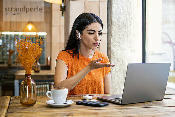 Junge Frau bläst Kuss bei Videoanruf über Laptop im Café zu