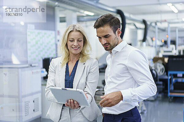 Lächelnde blonde Geschäftsfrau diskutiert mit einem männlichen Kollegen in der Fabrik über ein digitales Tablet