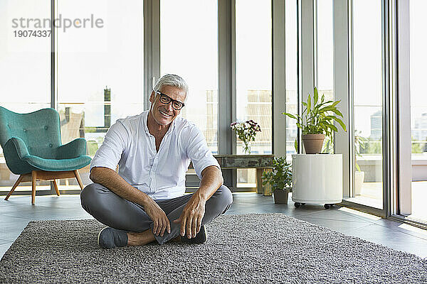 Porträt eines lächelnden reifen Mannes  der entspannt zu Hause auf dem Teppich sitzt