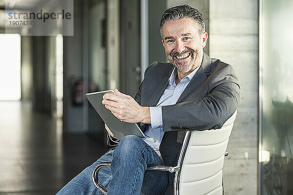 Porträt eines glücklichen  reifen Geschäftsmannes mit Tablet  der auf einem Stuhl im Büro sitzt