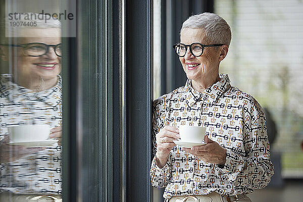 Lächelnde ältere Frau mit einer Tasse Kaffee  die aus dem Fenster schaut