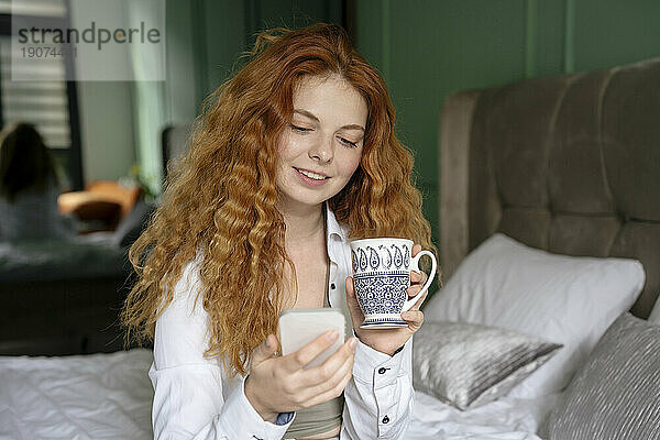 Junge Frau benutzt Smartphone und Teetasse im Bett