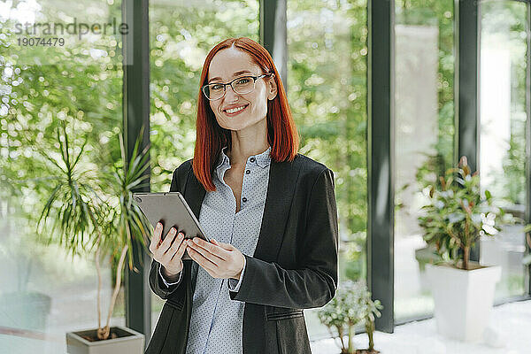 Glückliche rothaarige Geschäftsfrau steht mit Tablet-PC vor dem Fenster