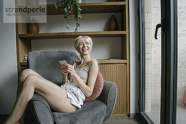 Glückliche Frau mit Smartphone entspannt sich zu Hause im Sessel
