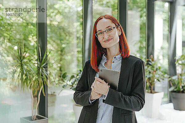 Lächelnde rothaarige Geschäftsfrau steht mit Tablet-PC im Büro