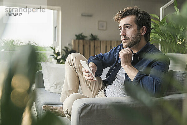 Junger Mann sitzt zu Hause auf der Couch und benutzt sein Smartphone