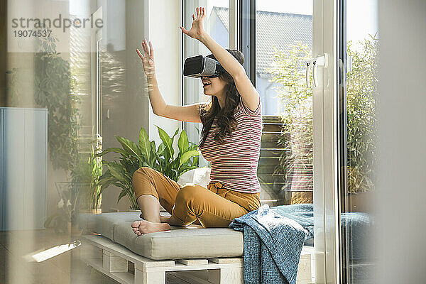 Junge Frau sitzt zu Hause am Fenster und trägt eine VR-Brille