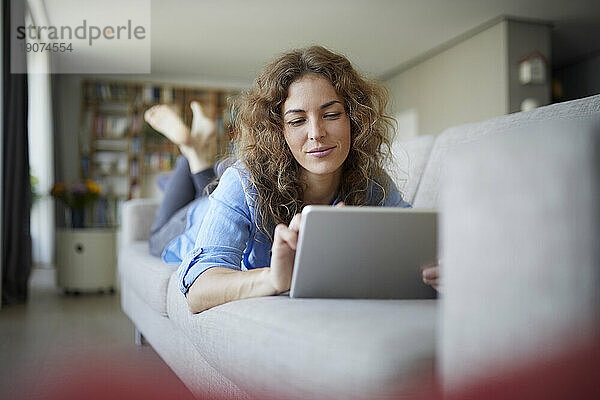 Schöne Frau benutzt digitales Tablet  während sie zu Hause auf dem Sofa liegt