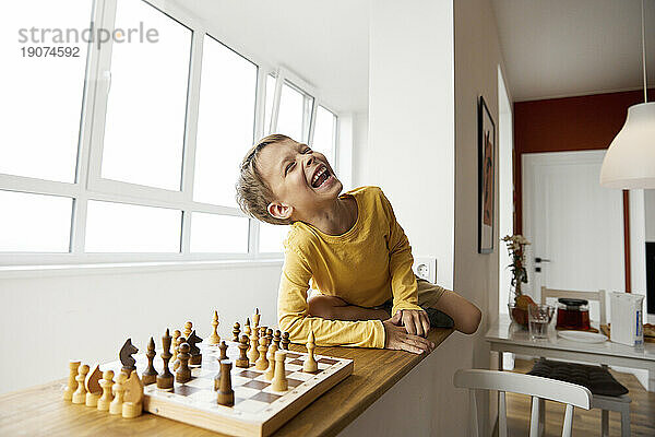Fröhlicher Junge spielt Schach und sitzt zu Hause auf der Fensterbank