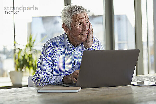 Porträt eines nachdenklichen älteren Mannes  der mit Laptop am Tisch sitzt und in die Ferne blickt