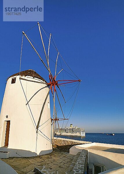 Berühmte Touristenattraktion von Mykonos  Kykladen  Griechenland. Traditionelle weiß getünchte Windmühle am Wasser und Kreuzfahrtschiff  das den Hafen verlässt  im Hintergrund. Sommer  Morgen  klarer blauer Himmel. Reiseziel  ikonische Ansicht. Diagonaler Aufriss