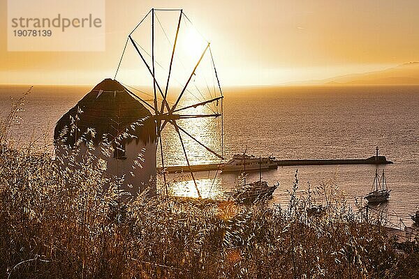 Goldener Sonnenuntergang über dem Meereshorizont in Mykonos  Kykladen  Griechenland. Berühmte traditionelle weiße Windmühle übersehen zivilen Hafen und Hafen auf heißen Sommerabend in hellem Sonnenlicht. Romantisches Bild  ikonische Reiseziele