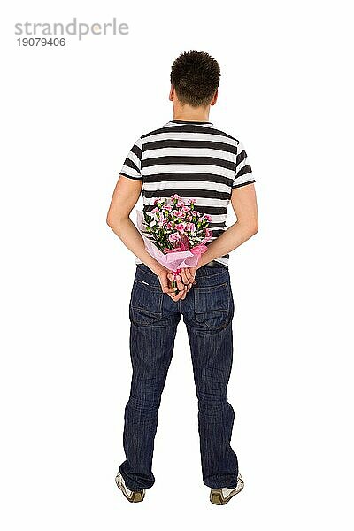 Lässiger junger Mann steht mit dem Rücken zur Kamera und versteckt einen Blumenstrauß hinter seinem Rücken  vor weißem Hintergrund
