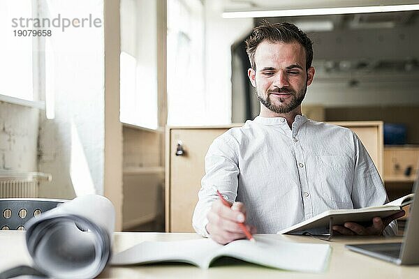 Lächelnder junger Mann  der auf einem Stuhl sitzt und im Büro arbeitet