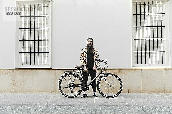 Bärtiger junger Mann steht mit Fahrrad an der Wand