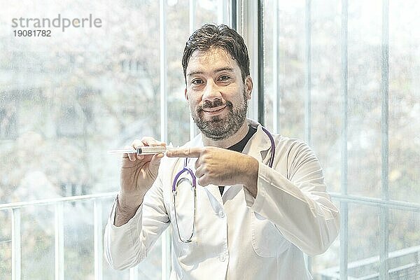 gutaussehender hispanischer Mann in Arztkleidung und Stethoskop in einer medizinischen Klinik  der in die Kamera lächelt  während er einen Impfstoff präsentiert