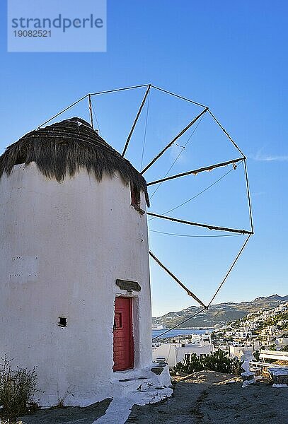 Berühmte Touristenattraktion von Mykonos  Kykladen  Griechenland. Traditionelle  weiß getünchte Windmühle auf einer Anhöhe über der Stadt Chora am Wasser. Sommer  Sonnenuntergang  blauer Himmel. Reiseziel  ikonische Ansicht. Vertikale Aufnahme