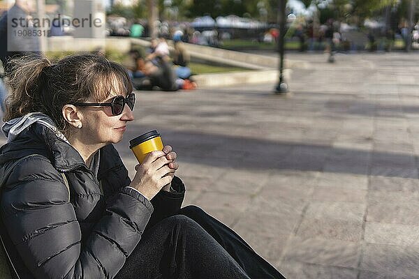 Ältere Frau sitzt auf einer Bank auf der Straße und trinkt entspannt und nachdenklich einen Kaffee bei Sonnenuntergang