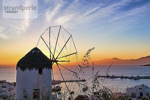 Berühmte traditionelle weiße Windmühle übersehen zivilen Hafen und Hafen von Mykonos  Kykladen  Griechenland bei Sonnenuntergang Himmel. Schöne Himmel  Sonne berühren Meer Horizont  Chora  Hauptort der Insel. Große bunte Wolken  selektiver Fokus  hohes Gras im Vordergrund