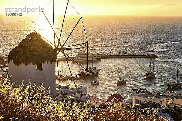 Goldener Sonnenuntergang über dem Meereshorizont in Mykonos  Kykladen  Griechenland. Berühmte traditionelle weiße Windmühle übersehen zivilen Hafen und Hafen auf heißen Sommerabend in hellem Sonnenlicht. Romantisches Bild  ikonische Reiseziele