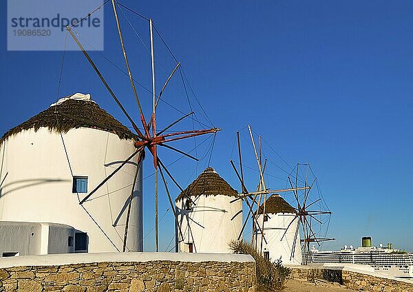 Berühmte Touristenattraktion von Mykonos  Kykladen  Griechenland. Drei traditionelle  weiß getünchte Windmühlen am Wasser. Sommer  blauer Himmel  Kreuzfahrtschiff beim Verlassen des Hafens im Hintergrund. Reiseziel  ikonische Ansicht