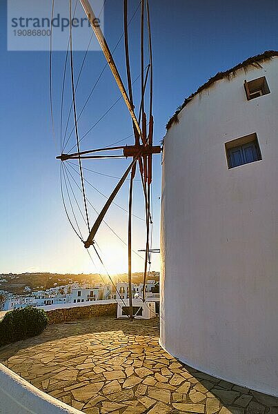 Berühmte Touristenattraktion von Mykonos  Kykladen  Griechenland. Traditionelle weiß getünchte Windmühle auf einem Hügel mit Blick auf Chora  Hauptort der Insel  gegen die Sonne. Sommer  Sonnenaufgang  blauer Himmel. Reiseziel  ikonische Ansicht. Vertikale seitliche Aufnahme