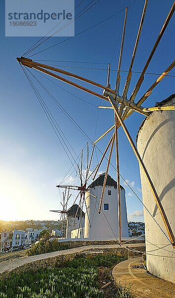 Berühmte Touristenattraktion von Mykonos  Kykladen  Griechenland. Drei traditionelle weiß getünchte Windmühlen auf einem Hügel mit Blick auf Chora  der Hauptort der Insel  gegen die Sonne. Sommer  Sonnenaufgang  blauer Himmel. Reiseziel  ikonische Ansicht. Vertikaler Aufriss