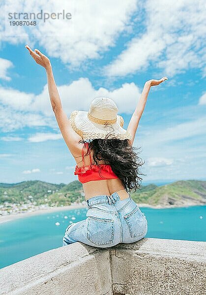 Lifestyle einer Frau im Urlaub sitzend mit ausgebreiteten Händen am Strand. Touristisches Mädchen mit Hut und ausgebreiteten Armen an einem Aussichtspunkt mit Blick auf den Strand