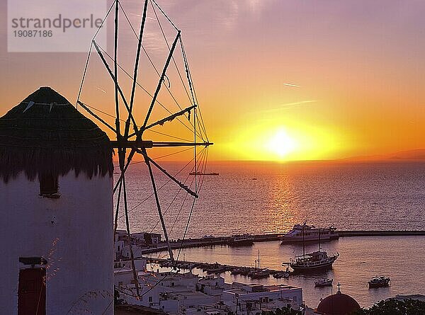 Berühmte traditionelle griechische Windmühle übersehen Hafen und Hafen von Mykonos  Kykladen  Griechenland bei Sonnenuntergang. Schöne Sonnenuntergang Himmel  Sonne berühren Meer Horizont  Hauptort der Insel in Sonnenuntergang Lichter und Schatten. Buntes Bild  selektiver Fokus  unterbelichtet
