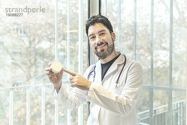 Gutaussehender hispanischer Mann in Arztkleidung und Stethoskop in einer medizinischen Klinik  der erstaunt in die Kamera lächelt  während er seine Hand präsentiert und mit dem Finger auf etwas zeigt