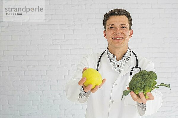 Lächelnder männlicher glücklicher Arzt bietet Apfel Brokkoli an