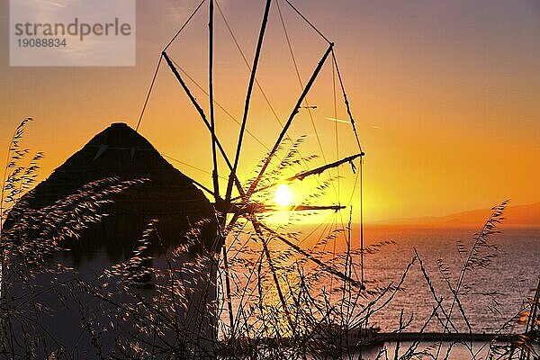 Silhouette des berühmten traditionellen griechischen Windmühle gegen bunten Sonnenuntergang Himmel  Meer und Sonne in Mykonos  Kykladen  Griechenland bei Sonnenuntergang. Sonne berühren Meer Horizont  hohes Gras im Vordergrund. Selektiver Fokus