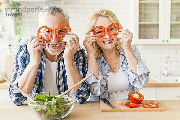 Lächelndes Porträt älteres Paar schaut durch rote Paprikascheibe