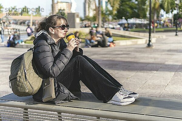 Ältere Frau sitzt auf einer Bank auf der Straße und trinkt entspannt und nachdenklich einen Kaffee bei Sonnenuntergang