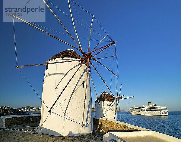 Berühmte Touristenattraktion von Mykonos  Kykladen  Griechenland. Traditionelle weiß getünchte Windmühlen am Wasser und ein Kreuzfahrtschiff  das den Hafen verlässt  im Hintergrund. Sommer  Morgen  klarer blauer Himmel. Reiseziel  ikonische Ansicht. Diagonaler Aufriss