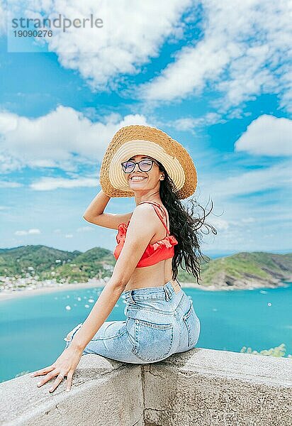 Porträt von Tourist Mädchen in einem Hut an einem Aussichtspunkt mit einem Strand im Hintergrund. Happy Tourist weiblich sitzt an einem Aussichtspunkt mit Strand im Hintergrund