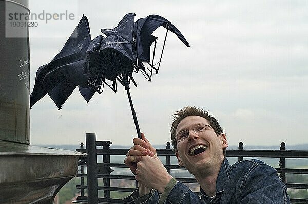 Spielerischer Umgang eines jungen Mannes beim Zusammenbrechen eines billigen Regenschirms durch eine Sturmböe auf einem Turm