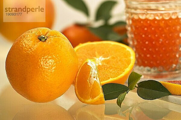 Vor einem Glas Saft liegt eine Apfelsine  eine halbierte Frucht und ein Stück Apfelsine sowie ein Zweig des Apfelsinenbaums. Im Hintergrund sind weitere Früchte und Zweige. Das Bild hat ein feines Bokeh