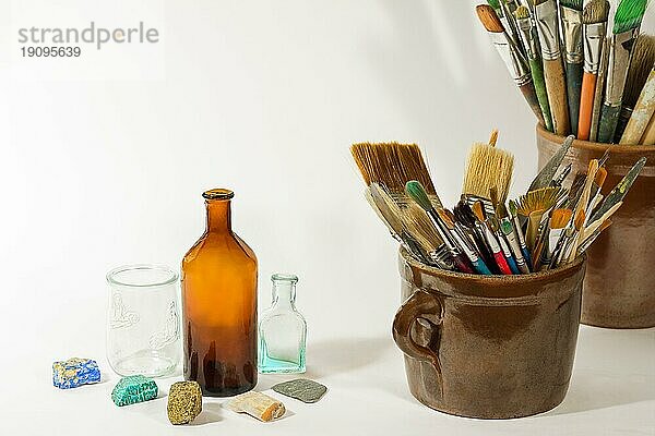 Künstlerische Pinsel  bunte schöne Steine  alte Flaschen und Malwerkzeuge in alten Keramiktöpfen auf weißem Hintergrund. Künstlerische Komposition
